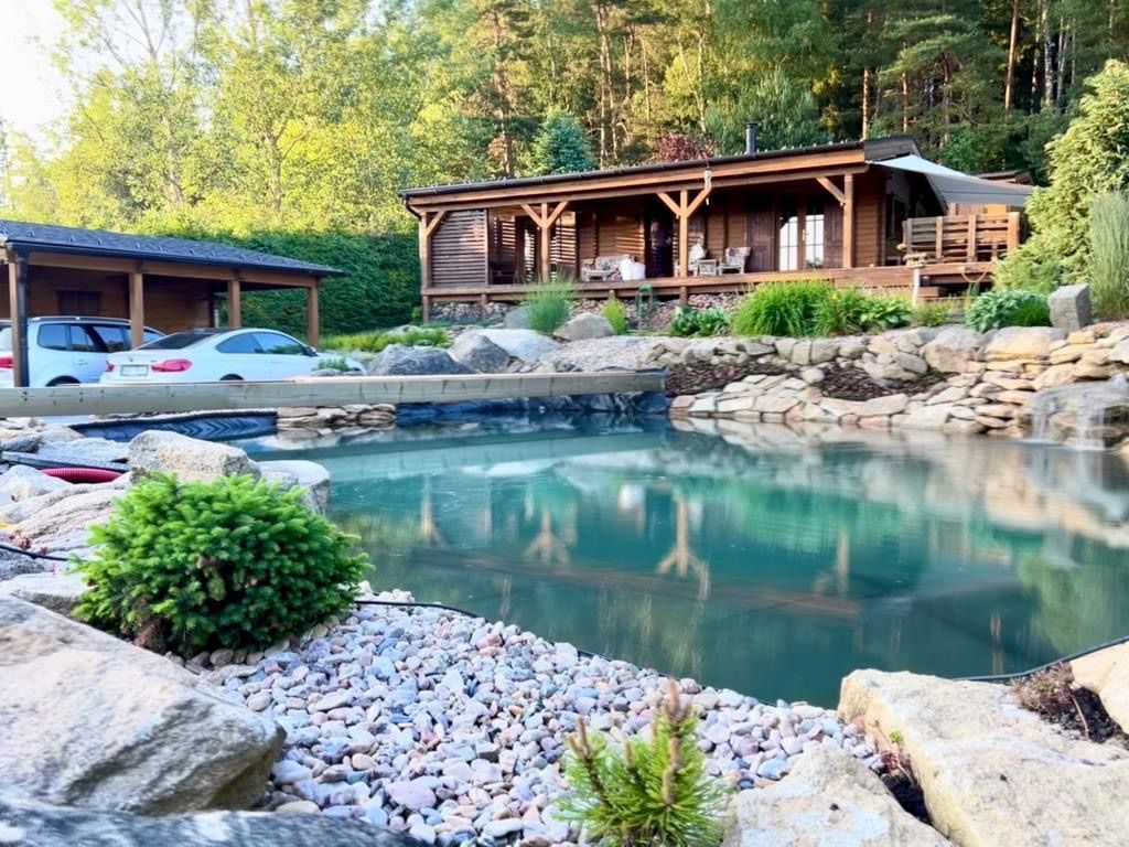 Wooden cabin with a pond, bridge, and a car shelter in a lush outdoor setting.