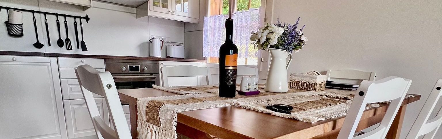 A kitchen with a wooden table set for dining. A bottle of wine and flowers are on the table.