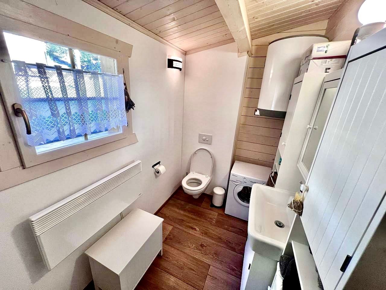 Small bathroom with a window, toilet, sink, and storage. Wooden floor and white walls.