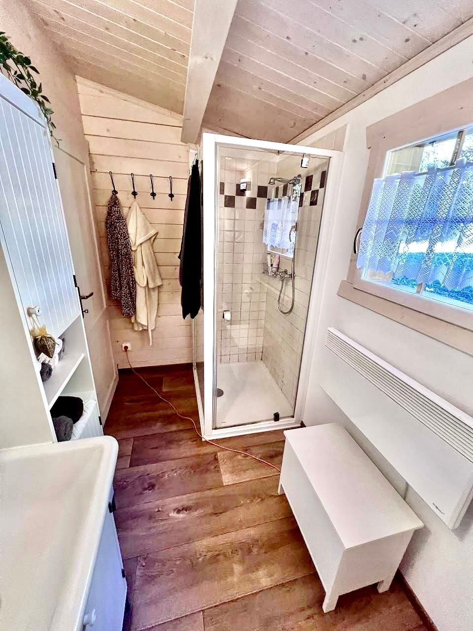 Small bathroom with shower, bench, sink, window, and wooden floor.