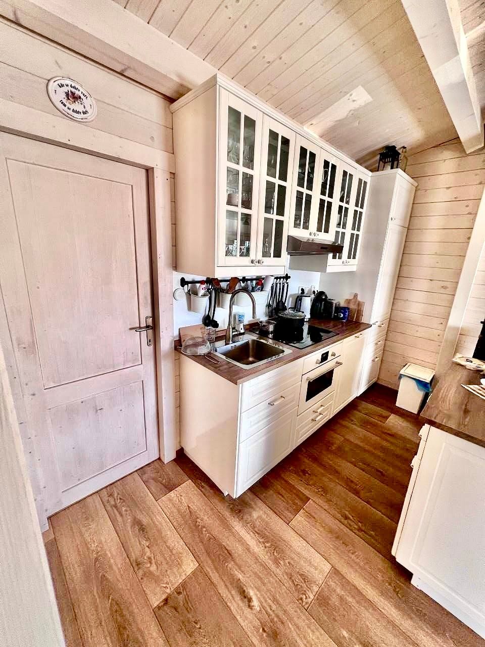 Small white kitchen with wood floors and cabinets, a sink, and appliances. A door is on the left.