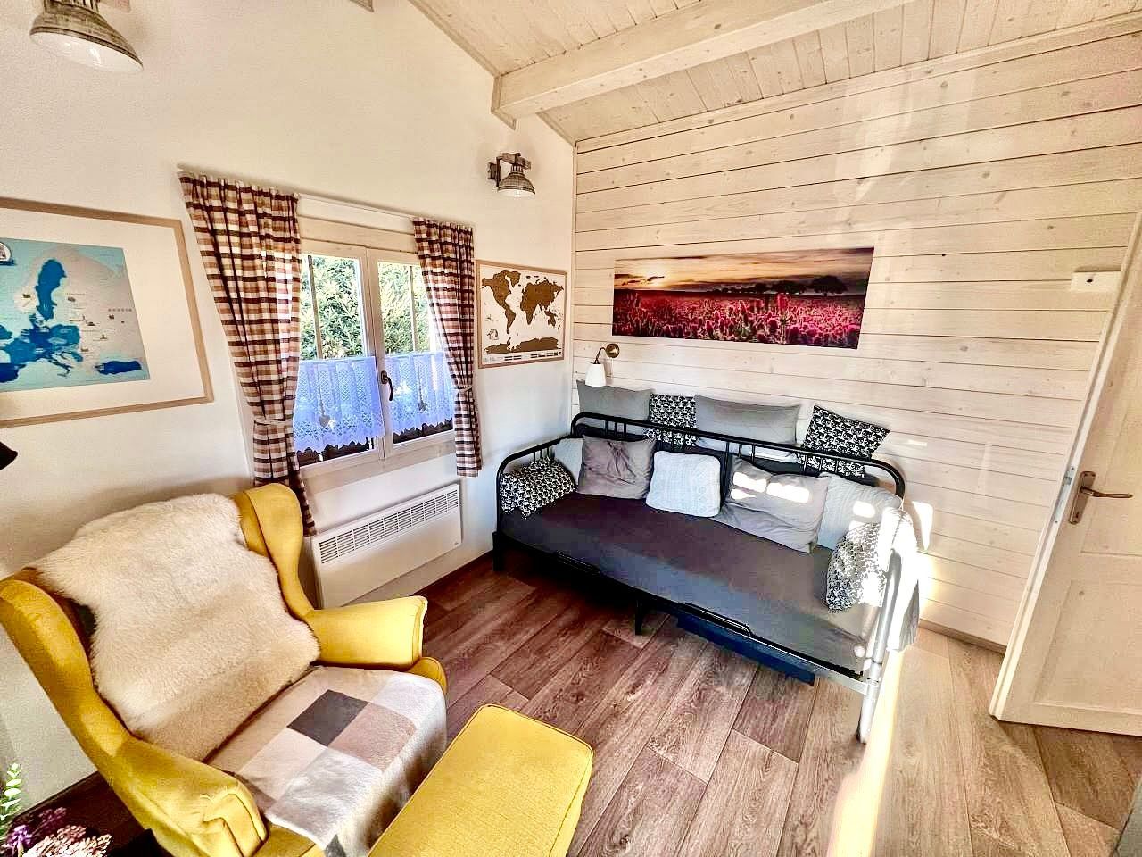 Cozy interior: yellow armchair, gray sofa, window with curtains, artwork, wooden floor, and white paneled walls.