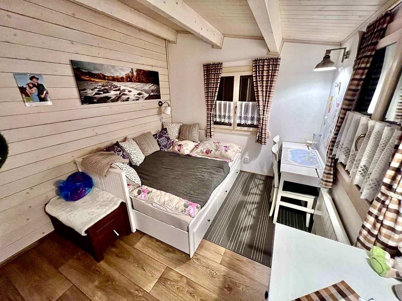 Cozy cabin bedroom with bed, window, desk, and chest. Walls are white with wood beams.
