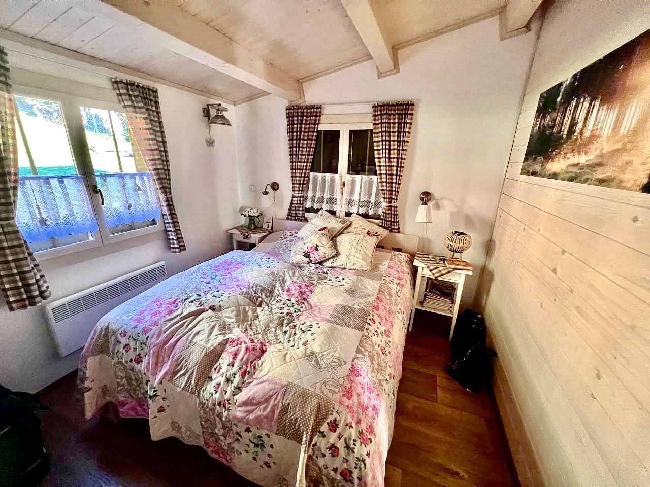 Cozy bedroom with patchwork quilt, plaid curtains, wood paneling, and a framed landscape photo.