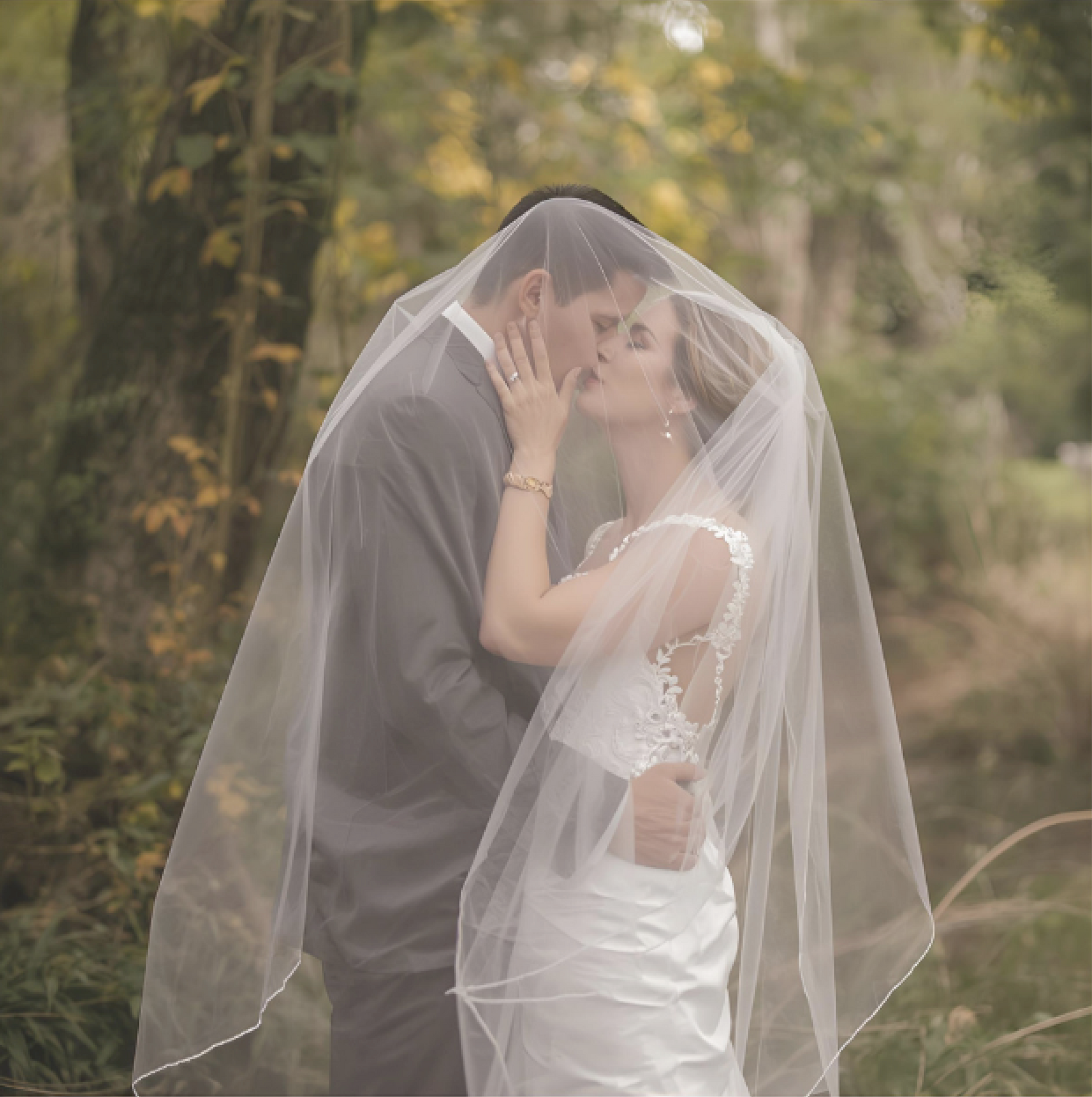 Nevěsta a ženich se líbají pod průsvitným závojem; venku se stromy v pozadí.