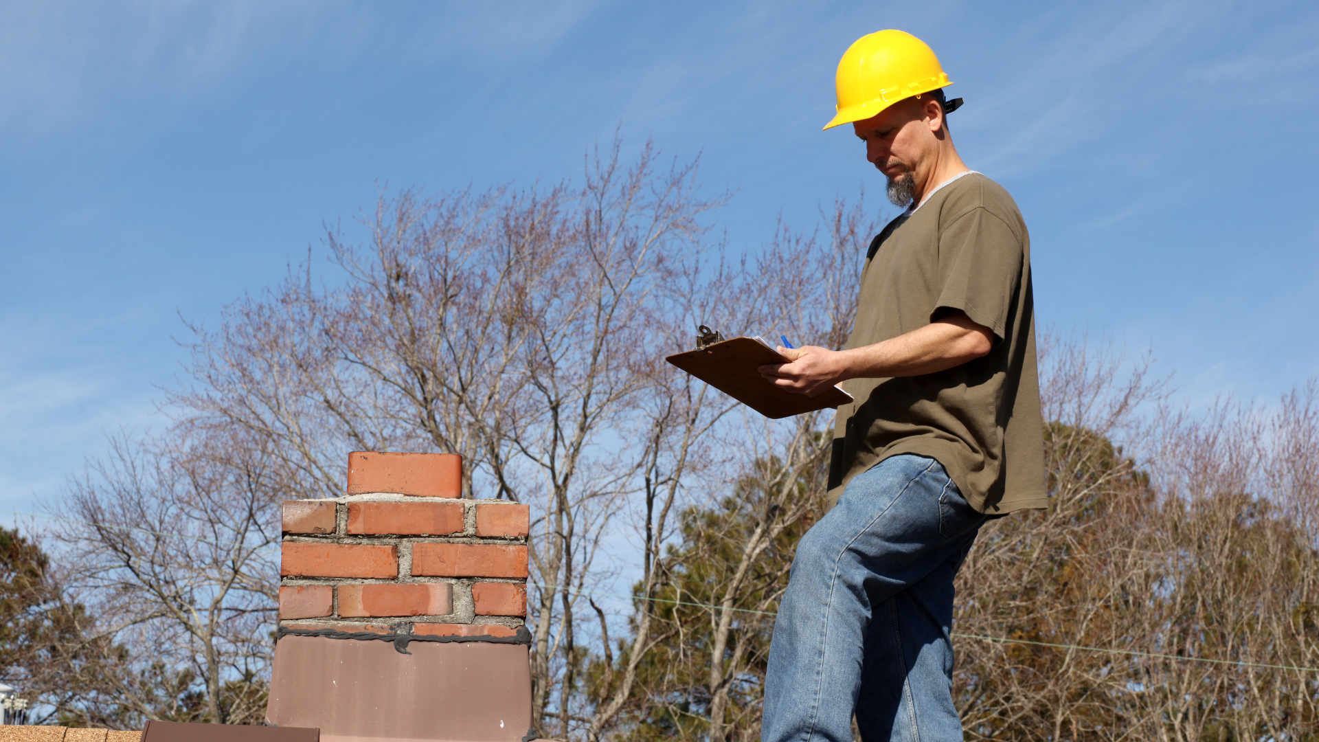 A man wearing a hard hat is standing on a roof looking at a clipboard.