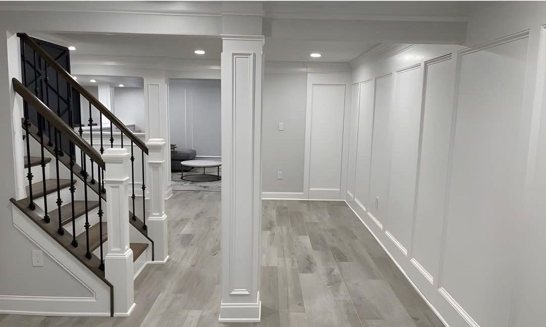 Newly renovated basement with a staircase, wooden floors, and white paneled walls. A central column stands in the hallway.