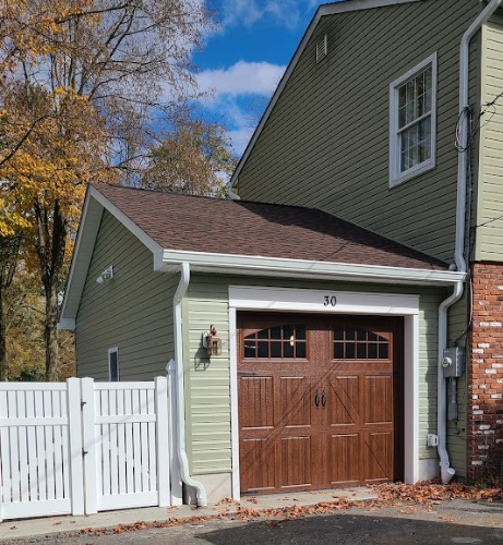 Green-sided garage with brown roof and door, white fence in front, brown leaves, and blue sky visible.