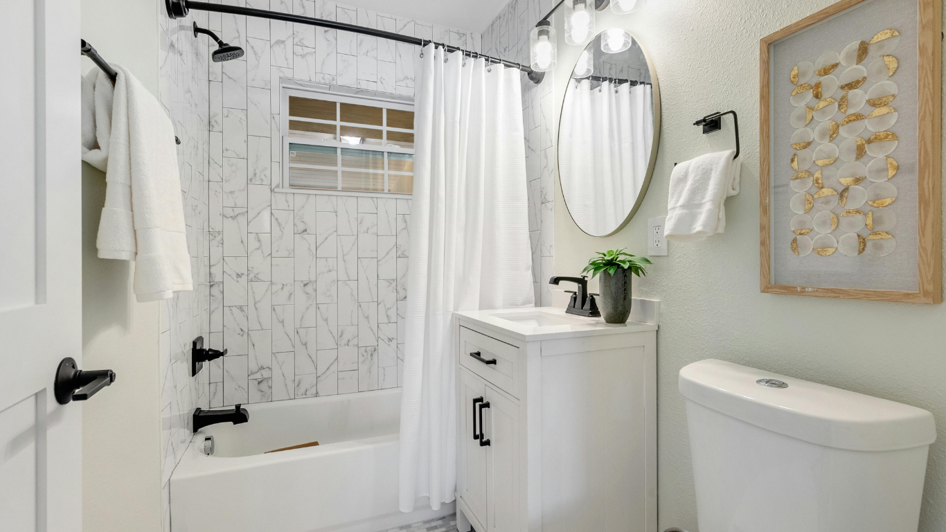 Small, bright bathroom with white fixtures and marble tile; black accents and a gold-framed art piece.