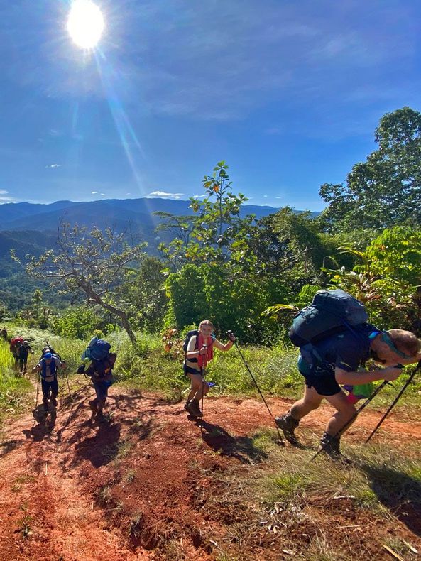 Image depicts a Kokoda Expedition group hiking up the final peak into Nauro Village along the Kokoda Track. The earth is orange in colour and the trekkers are using poles and carrying backpacks.