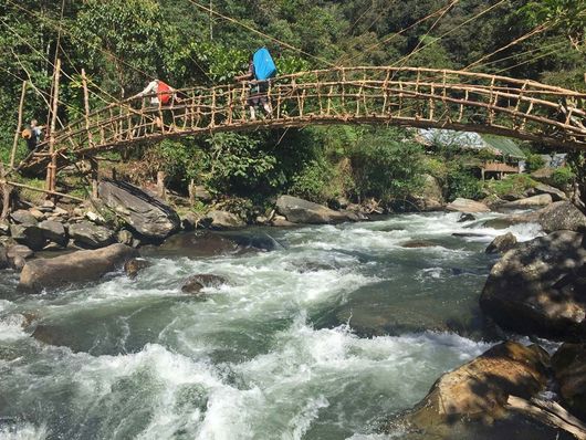 Image depicts a major river along the Kokoda Track with a vine bridge going across it and two people walking across the bridge. 