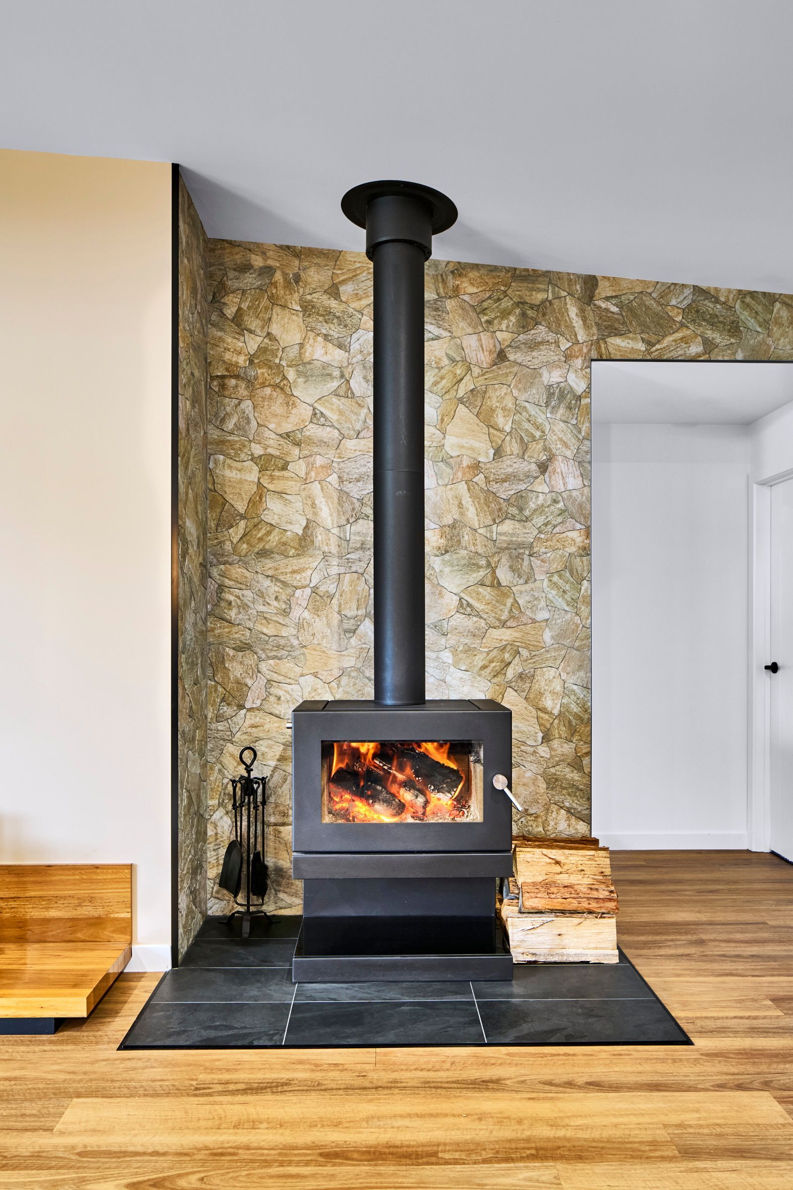Black wood-burning stove with lit fire, black chimney, and stone accent wall in a room with wood flooring.