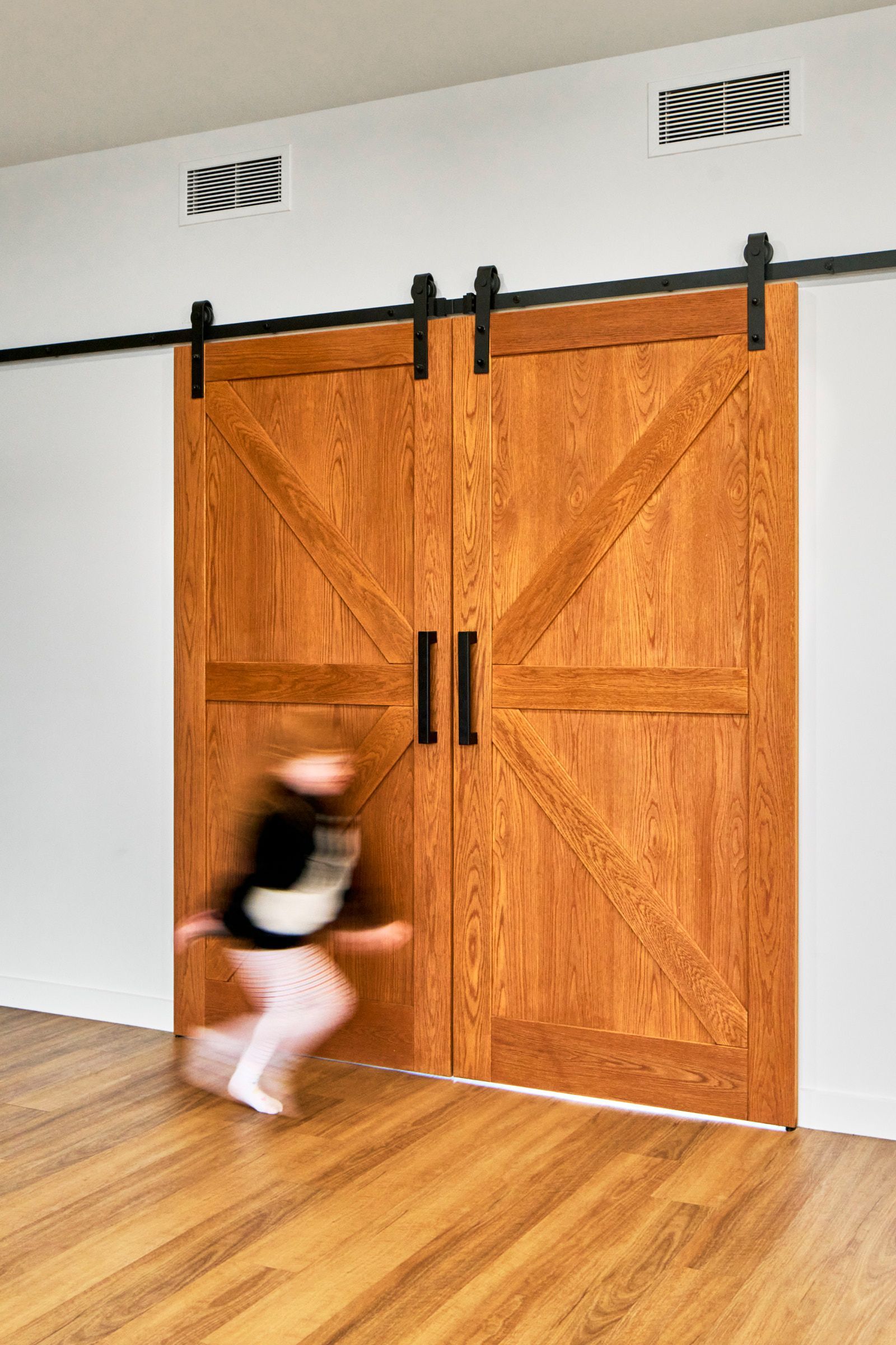 A child in motion runs past large, wooden barn doors on a hardwood floor; the doors are mounted on a black track.