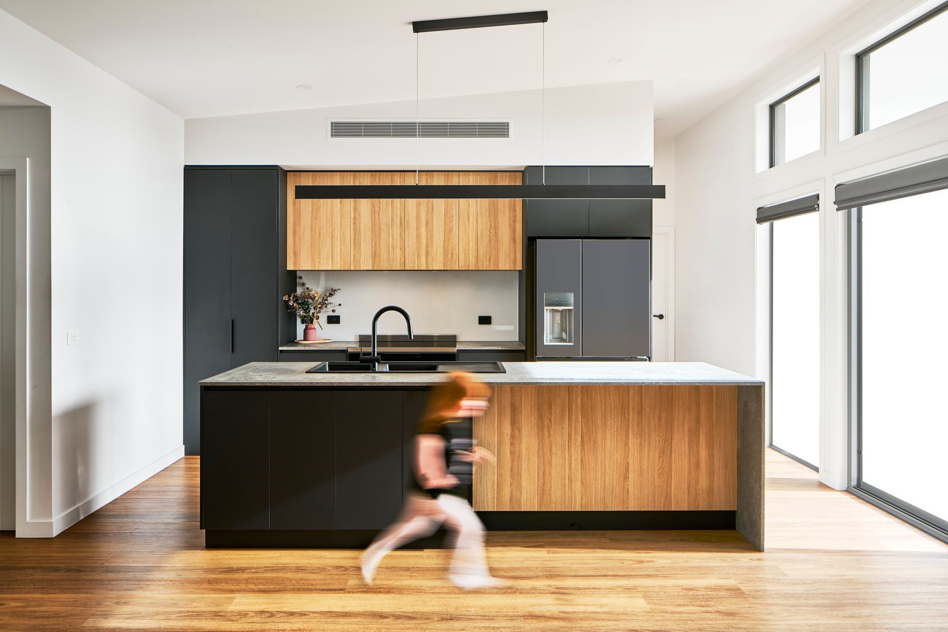Modern kitchen with black and wood cabinets, a large island, and a child running across the wooden floor.