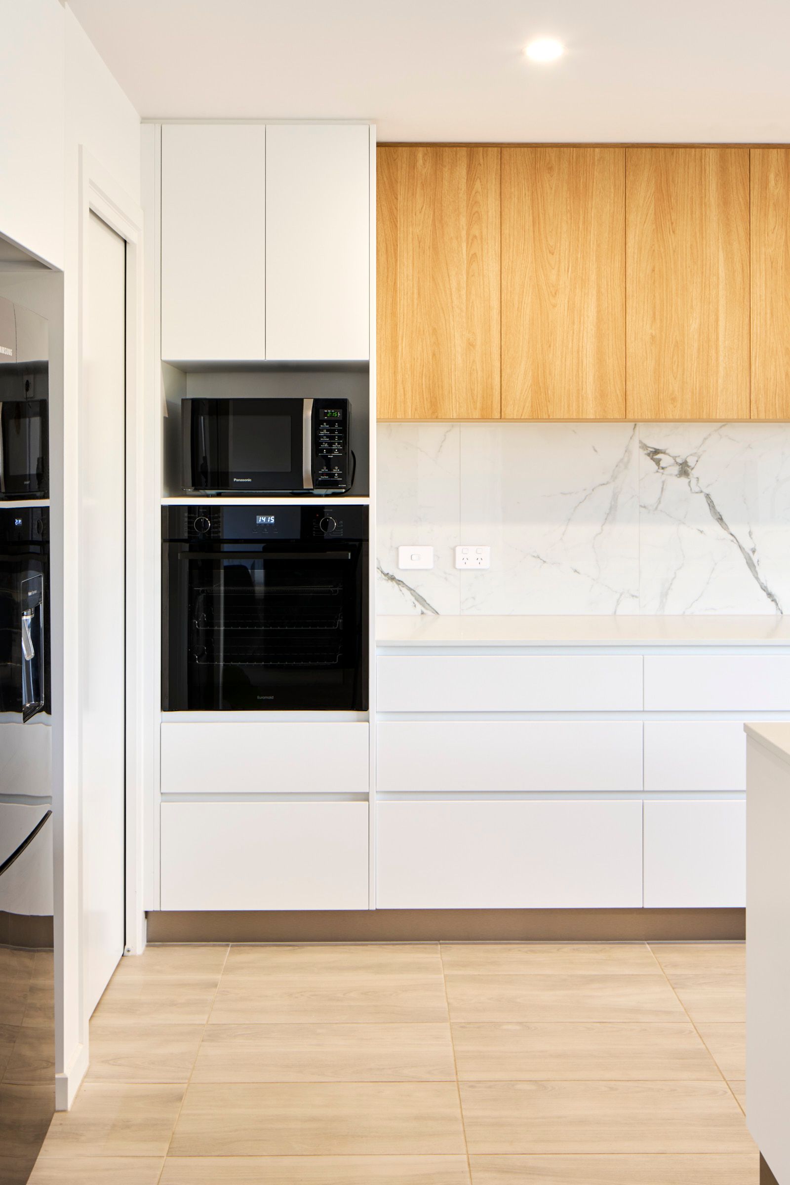 Modern kitchen with white and light wood cabinetry. Includes a built-in microwave and oven, with marble backsplash.
