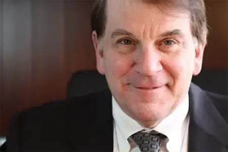 Smiling older man in a suit, light skin, brown hair, indoors near a dark wall.