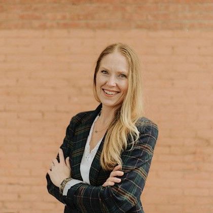 A woman in a plaid jacket is standing in front of a brick wall with her arms crossed.