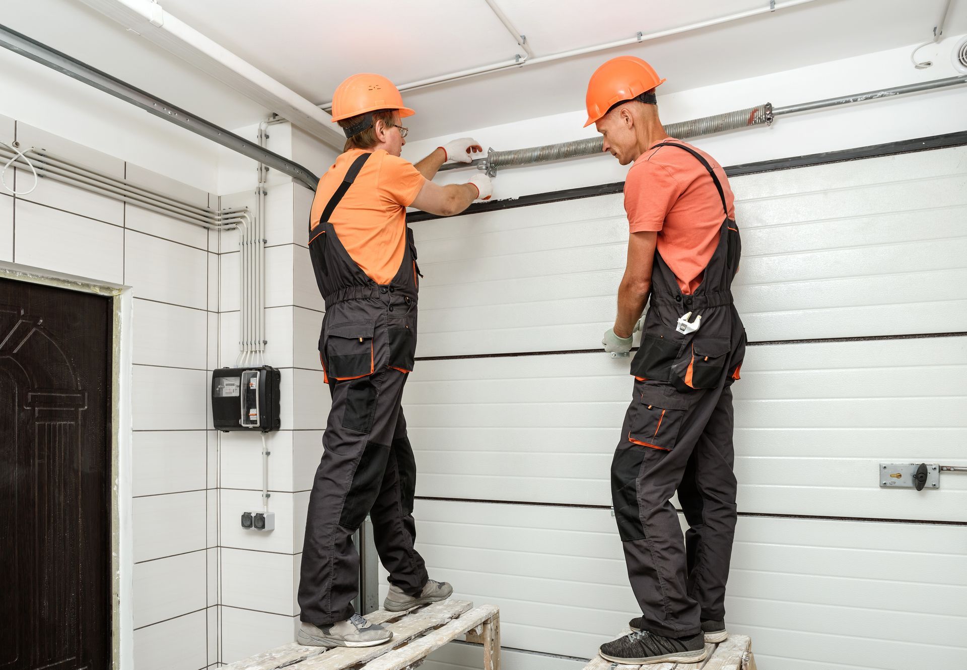 Two workers in orange uniforms are performing garage door service, installing lift gates in a door.
