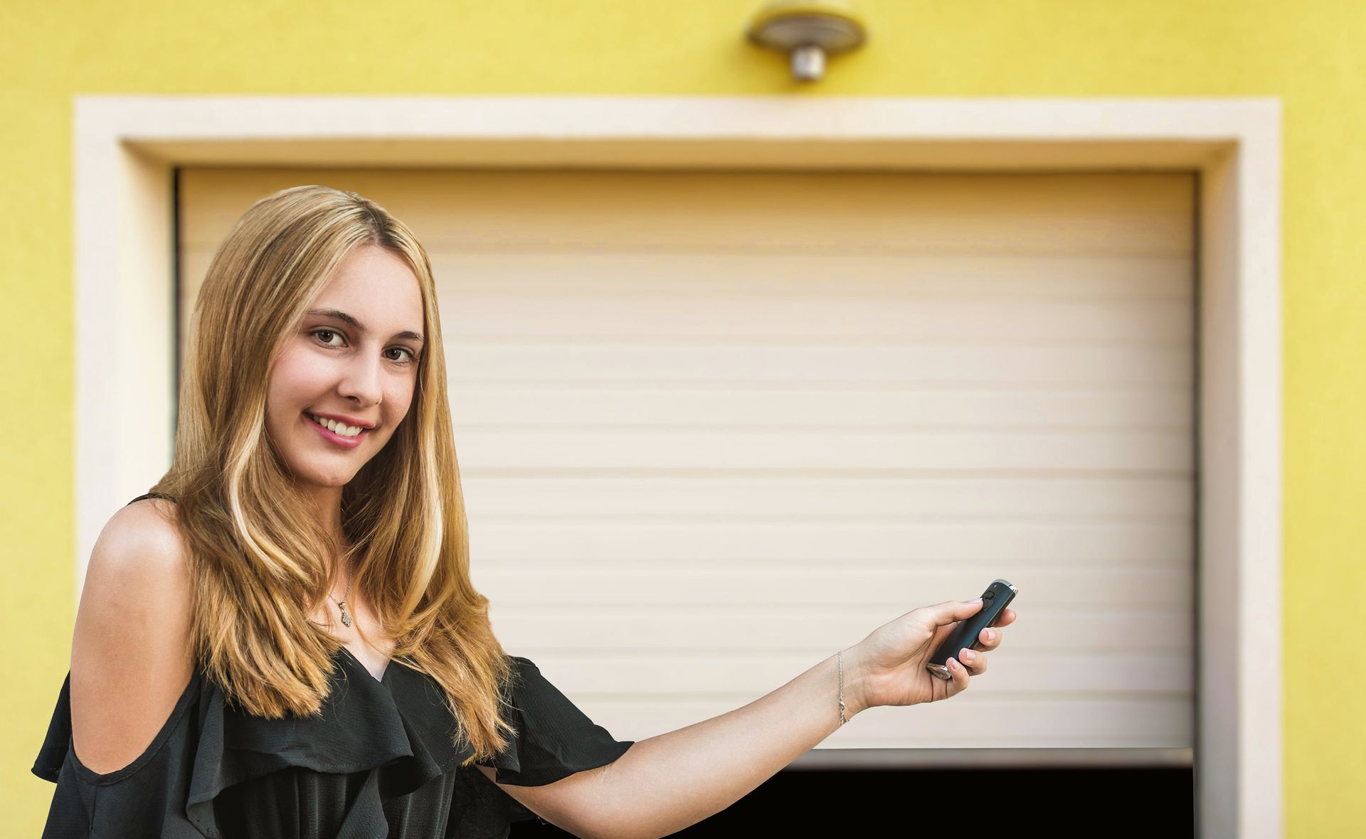 Blonde woman smiling, holding a garage door opener, standing in front of a beige garage door on a yellow wall.
