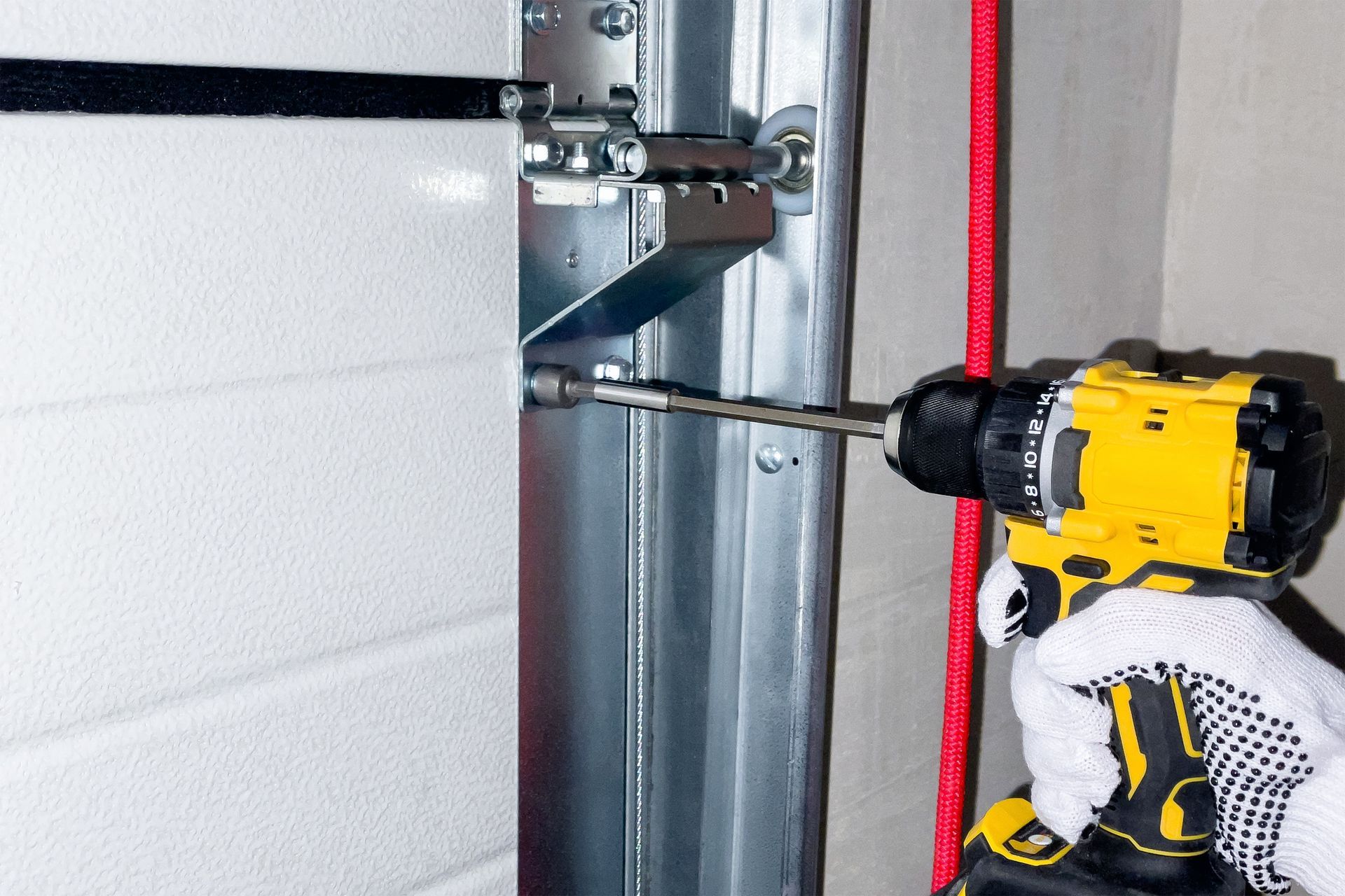 A person uses a power drill to install a bolt on a metal garage door frame, with a red rope visible nearby.