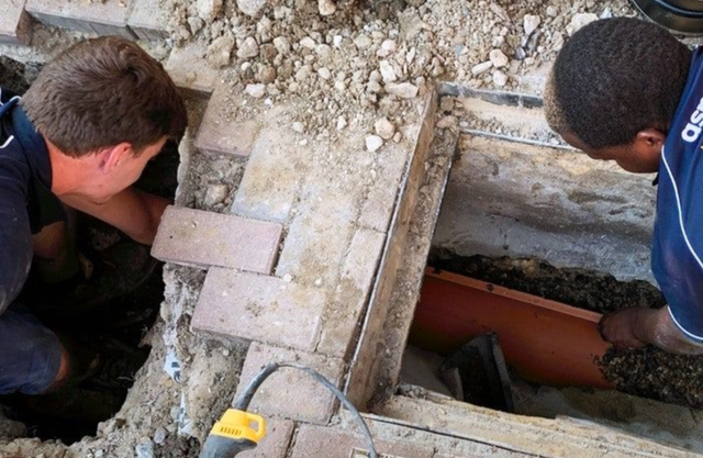 Two workers inspecting a concrete pit with exposed pipes and rubble on a construction site