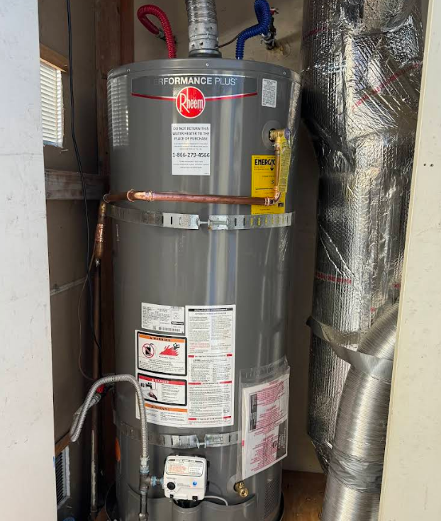 Gray water heater in a utility closet, with pipes and insulation around it.