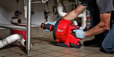 Plumber using a red drain-cleaning machine under a sink in a tiled utility room