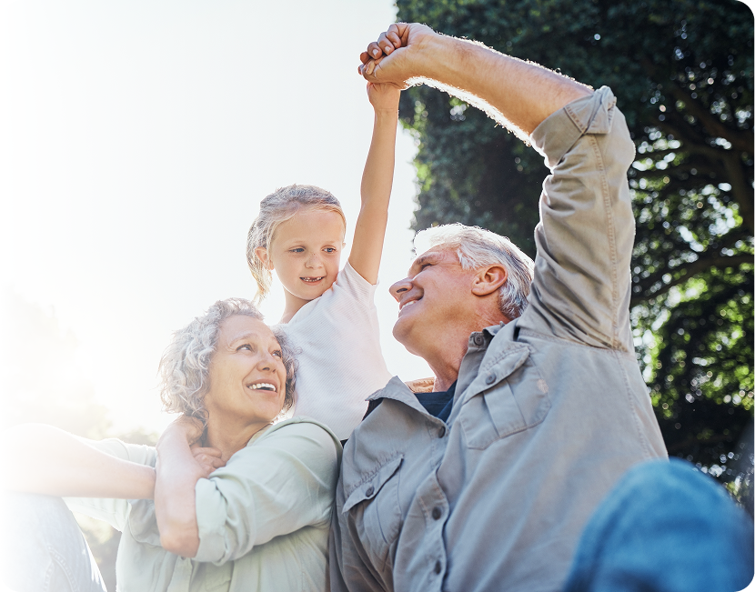 Grandparents lifting granddaughter up, arm raised; outside, happy smiles.