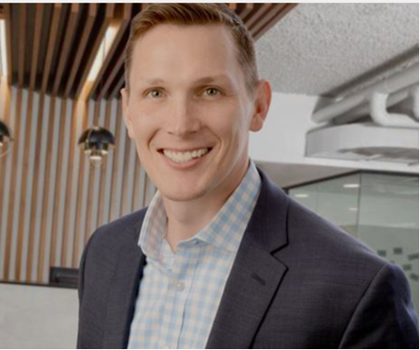 Founder, Drew McDermitt, smiling at the camera in a gray suit with a white and blue checkered button up shirt underneath