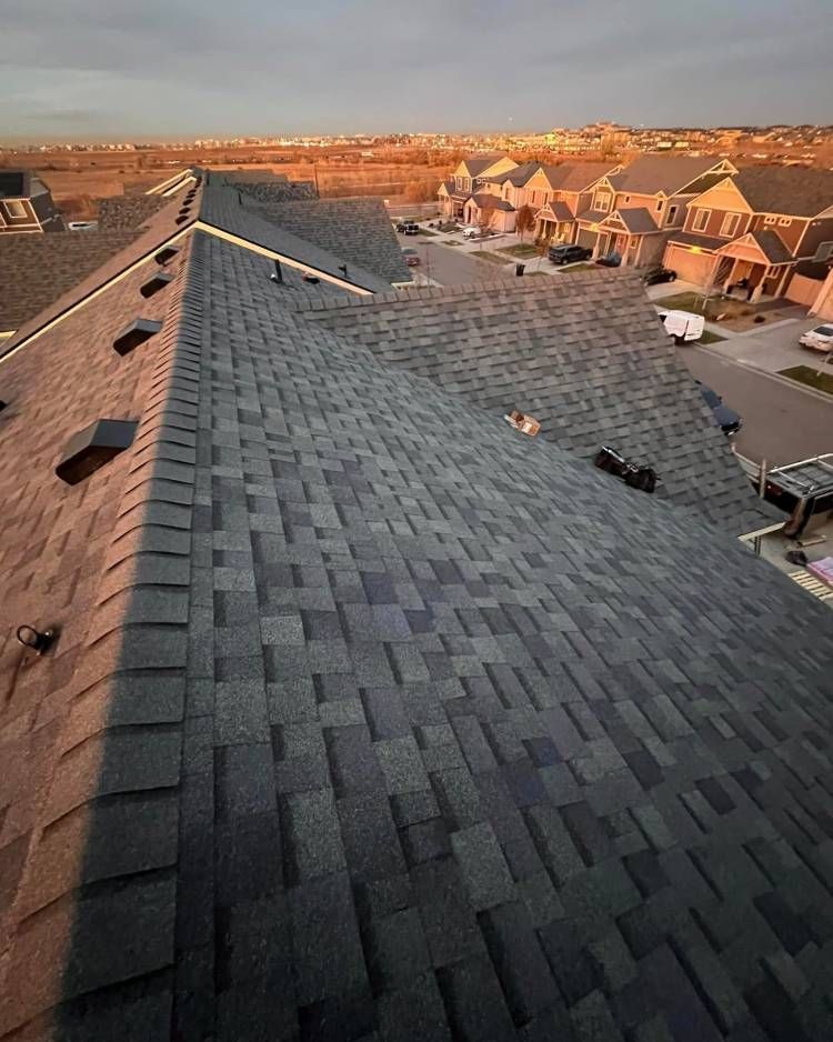 Overhead view of a dark shingle roof at sunset, overlooking a suburban neighborhood with houses.