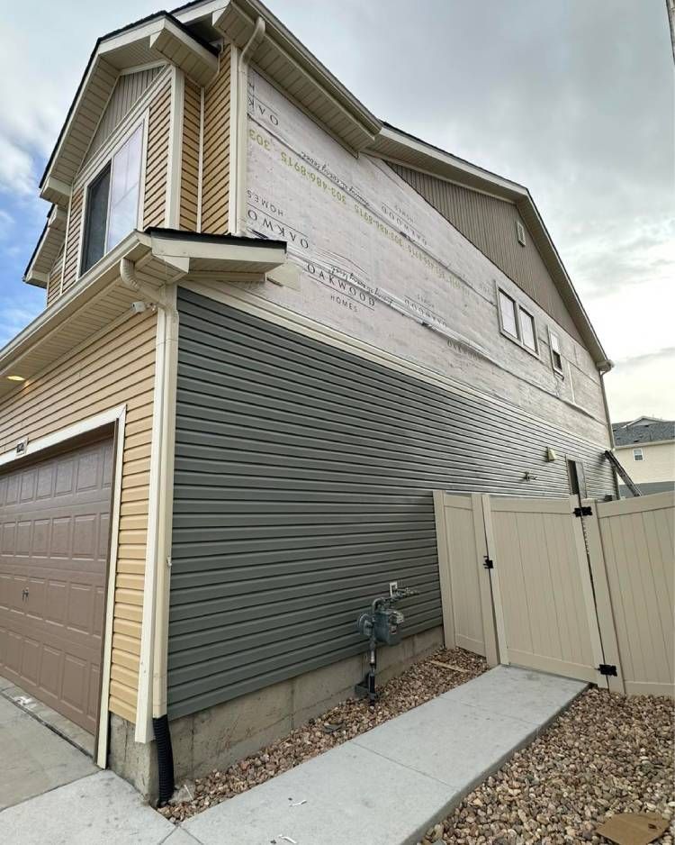 Two-story house with tan and gray siding, tan garage door, and a white fence. A concrete walkway leads to the fence.