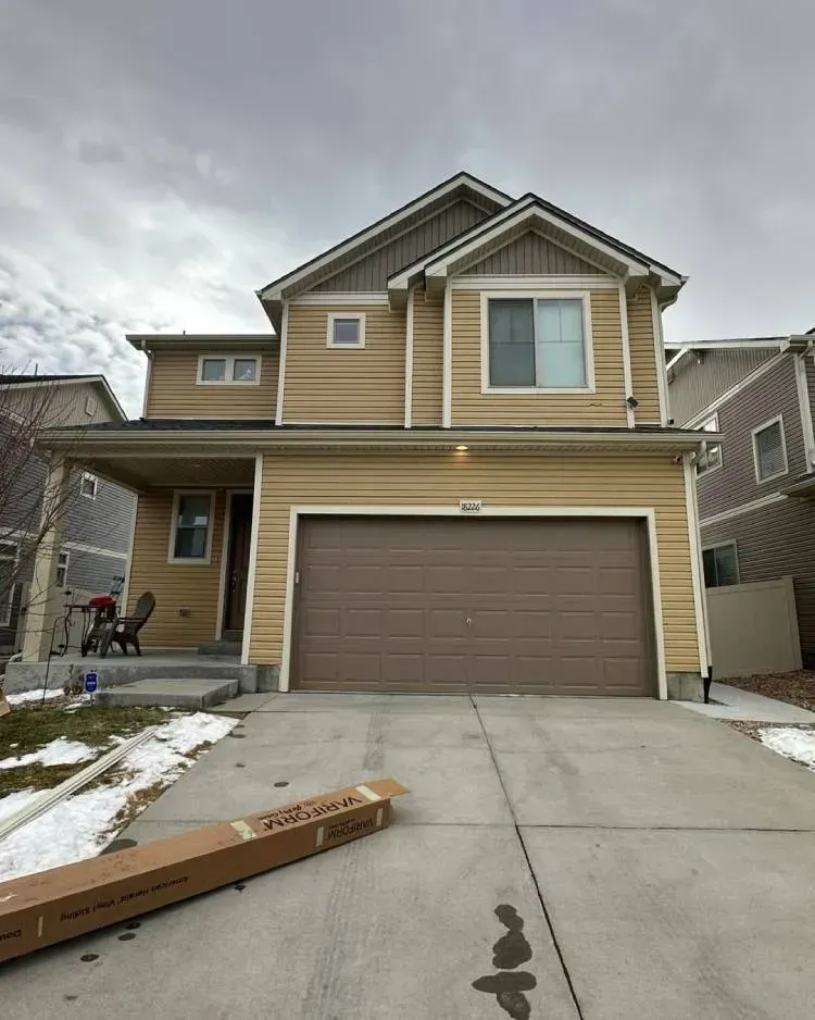 Two-story tan house with a brown garage door and a driveway. Snow on the ground; overcast sky.