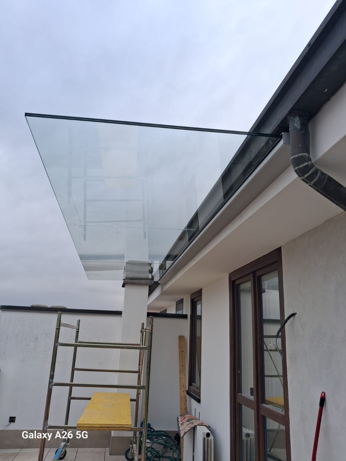 Pensilina in vetro fissata a un edificio bianco. Sono visibili un'impalcatura e una grondaia. Cielo nuvoloso.