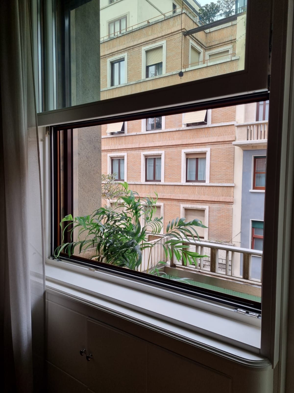 Vista attraverso una finestra di un condominio. Sono visibili edifici e vegetazione.