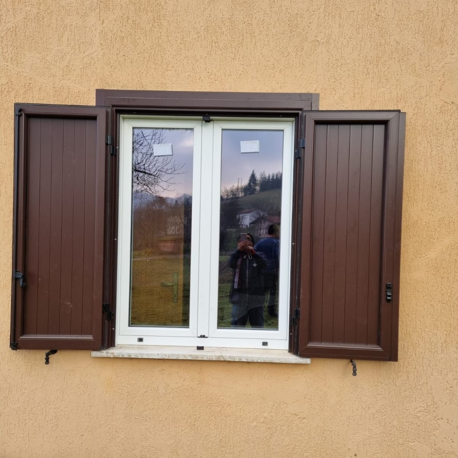 Finestra con persiane marroni su parete in stucco giallo; il riflesso mostra delle persone.