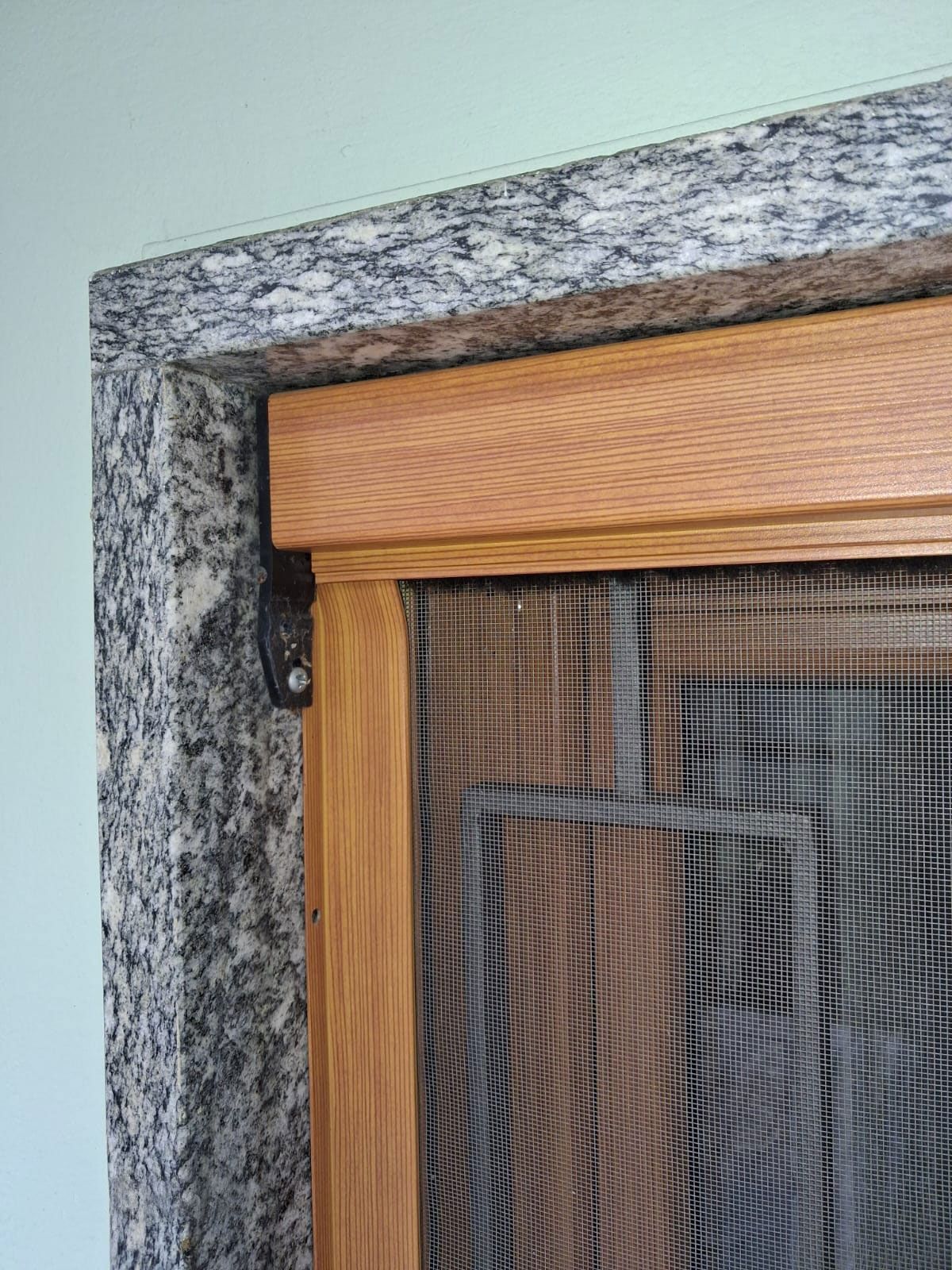 Primo piano di una porta a zanzariera incastonata in un telaio di granito. La porta è in legno con rete nera e si vede un angolo aperto.