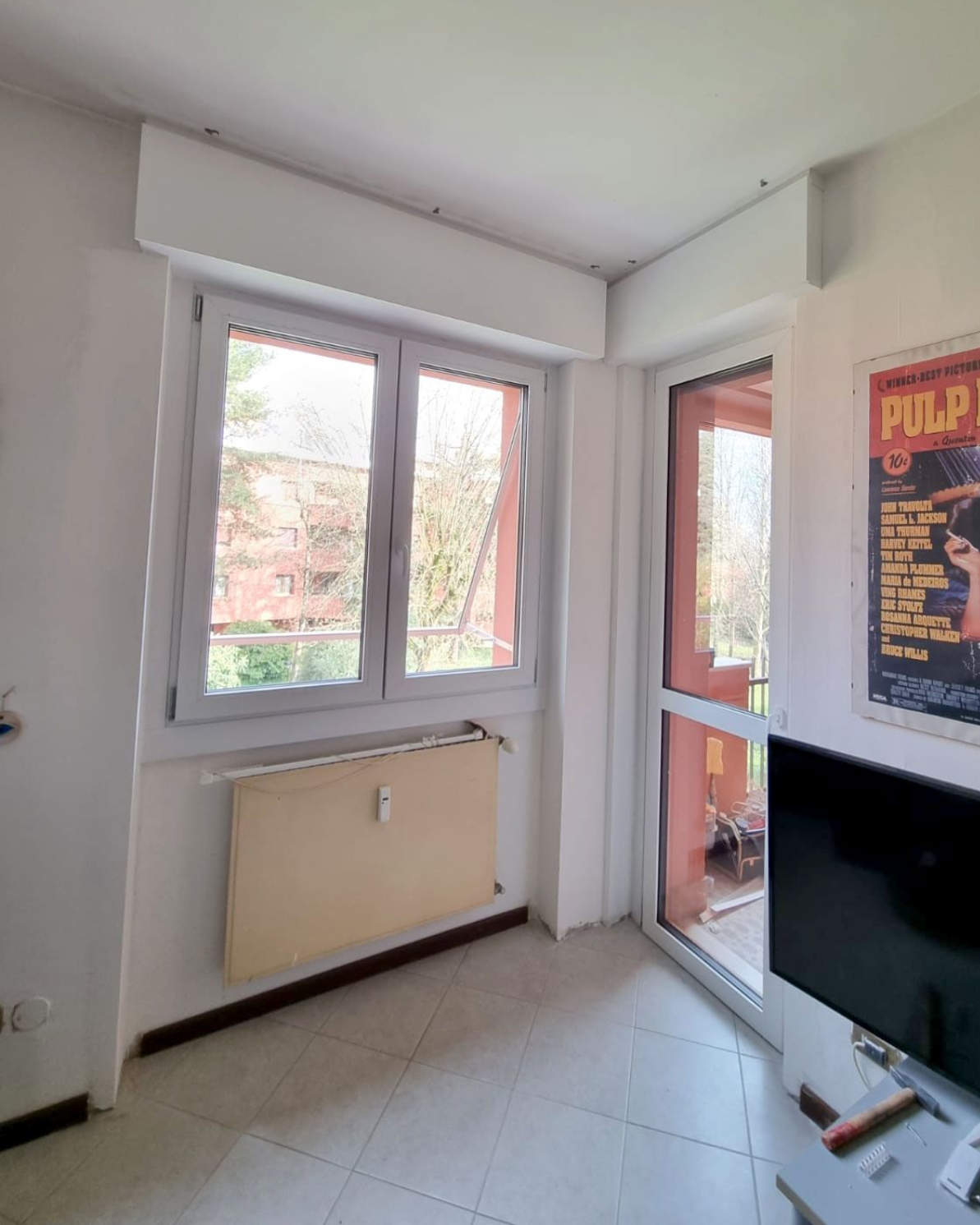 Vista interna con finestra, porta sul balcone e poster. Pareti chiare e pavimento piastrellato.