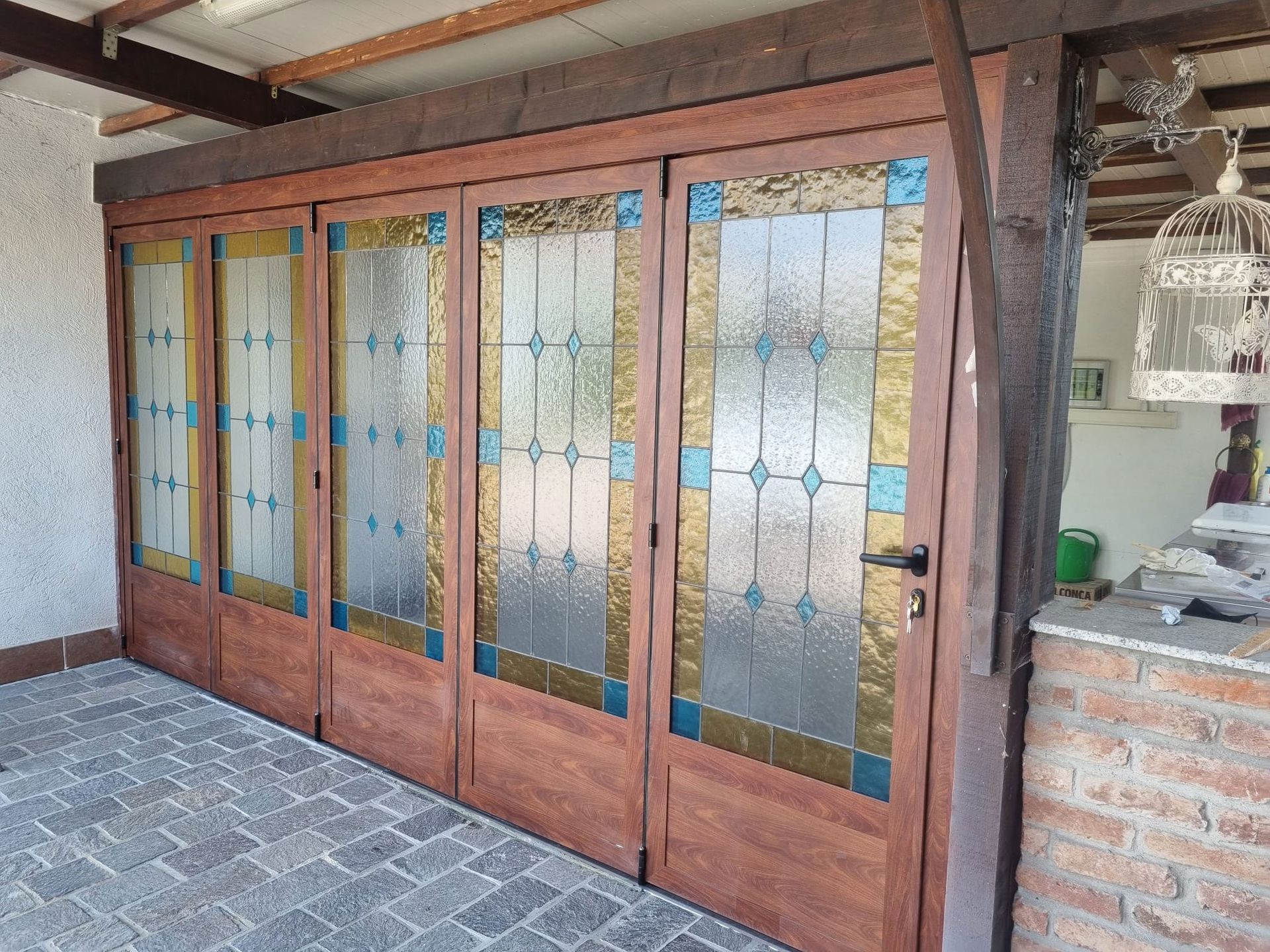 Quattro porte pieghevoli in legno con pannelli in vetro colorato. Situata su un patio in mattoni, sotto una tettoia in legno.