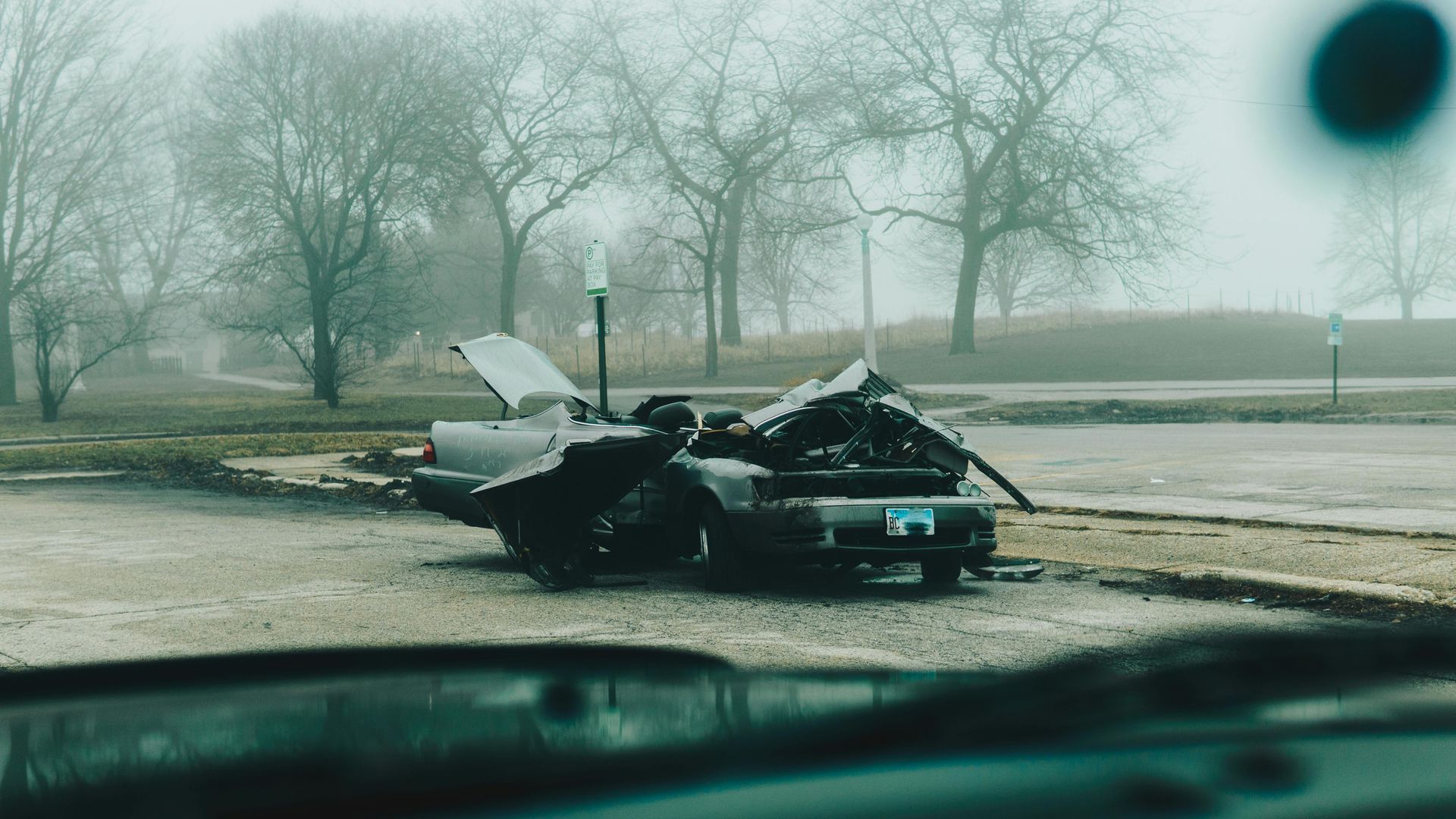 Severely damaged black car on a foggy road, with debris around it and a dark smudge on the windshield