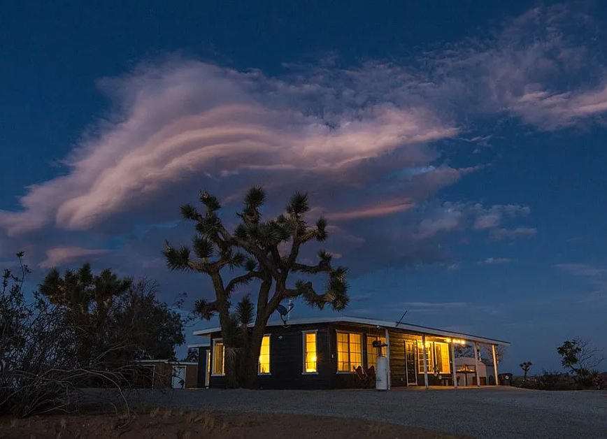 Desert cabin at dusk, illuminated windows, Joshua tree, dramatic cloud formations in a dark blue sky.