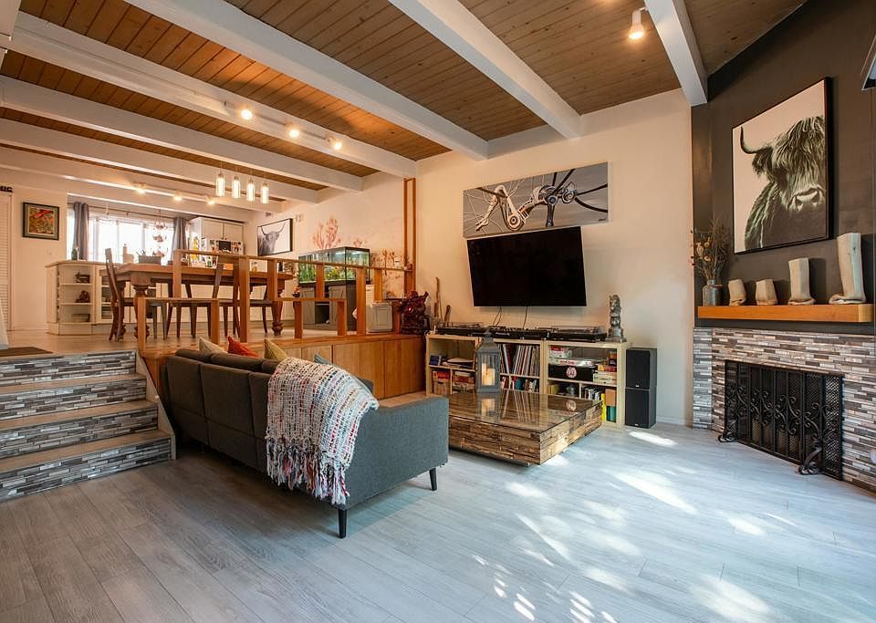 Living room with gray sofa, fireplace, TV, dining area, and wood beam ceiling.