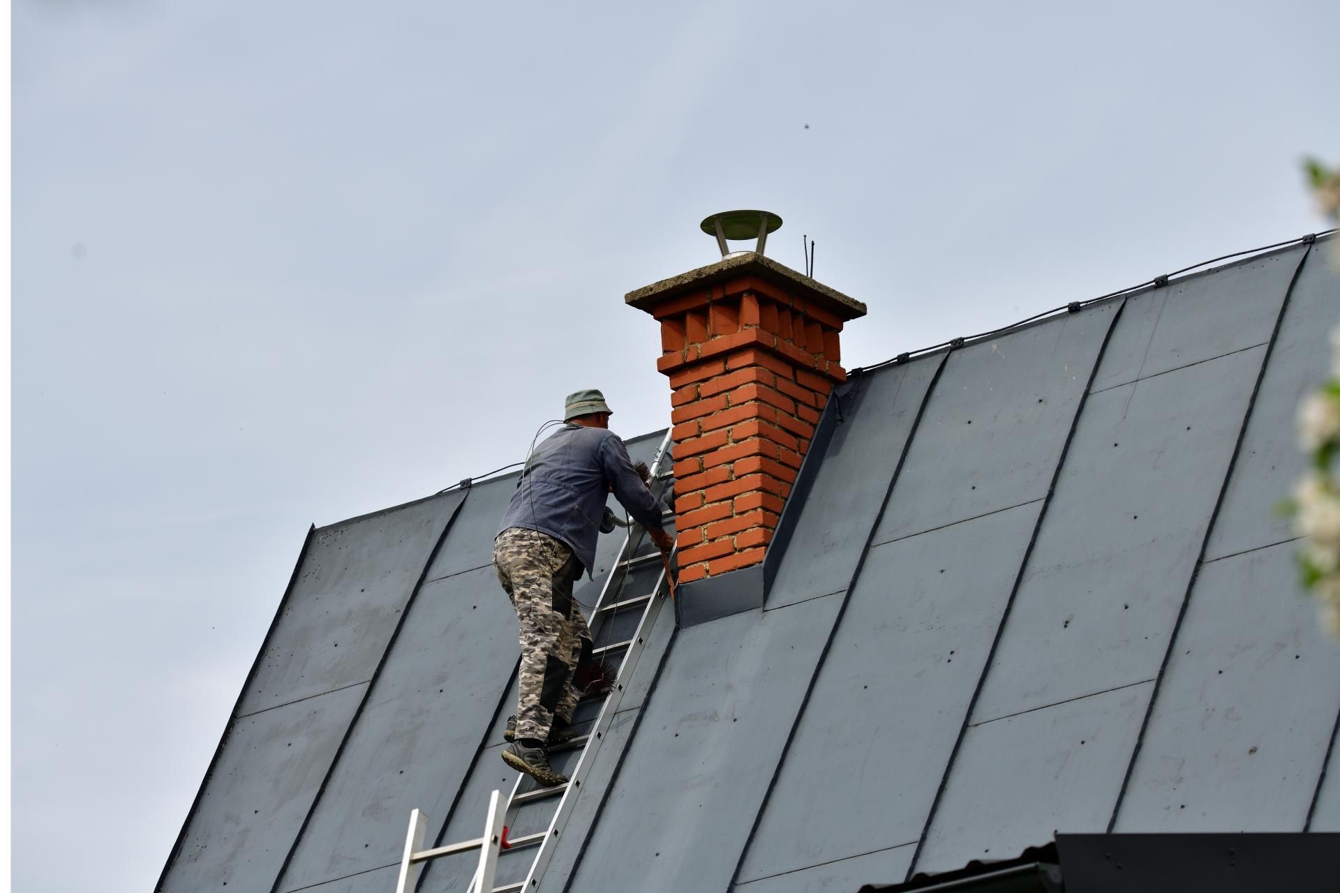 Person on a ladder, working on a chimney of a gray roof, overcast sky.