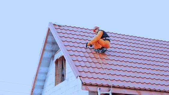 Roofer in orange jumpsuit works on a red metal roof, secured by a safety harness and rope.