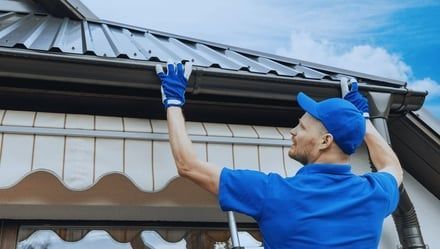 Man in blue cleaning gutters on a house, reaching up.