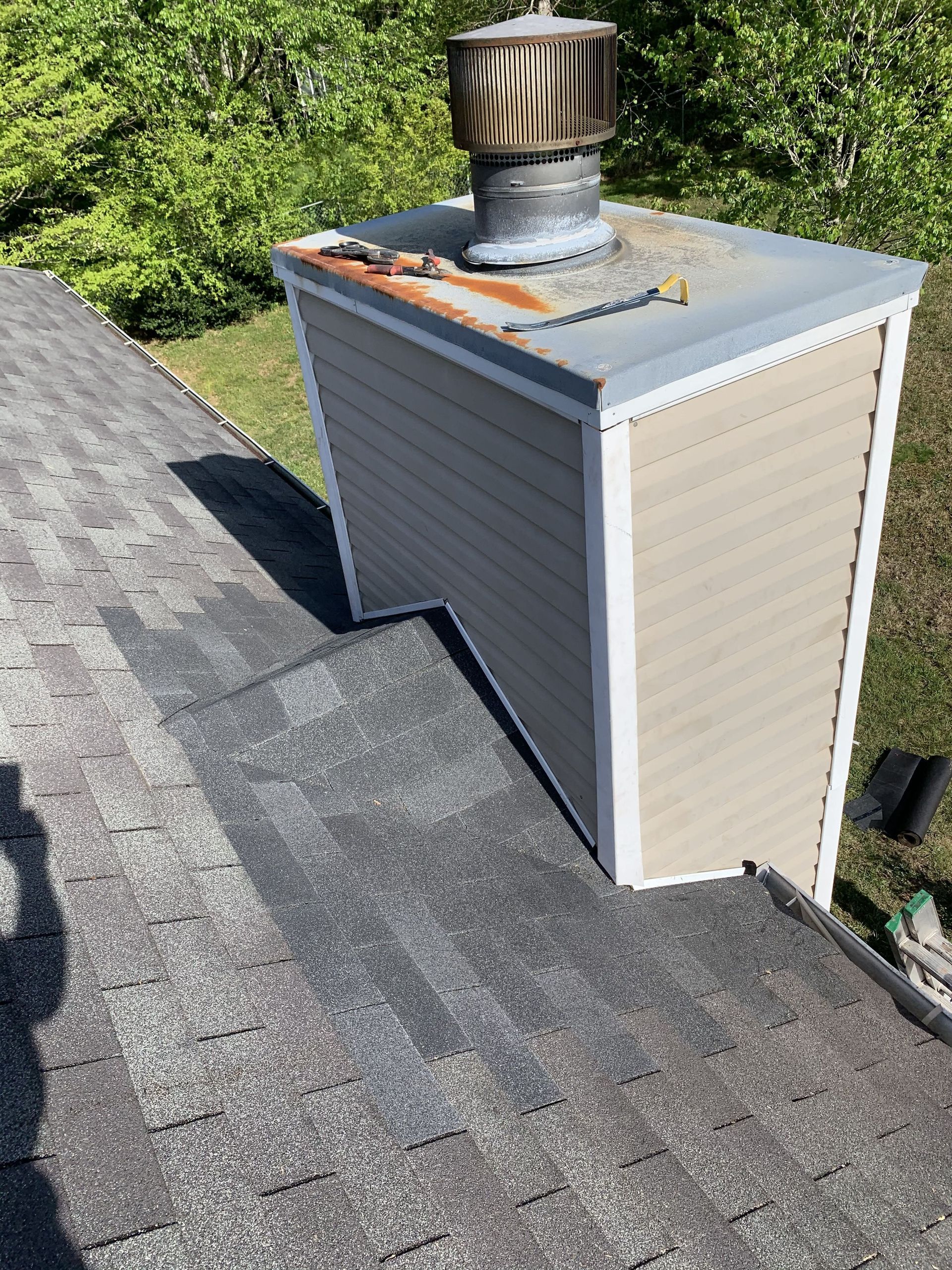 Chimney on a gray asphalt shingle roof. Tan siding, silver metal cap, and surrounding green trees are visible.