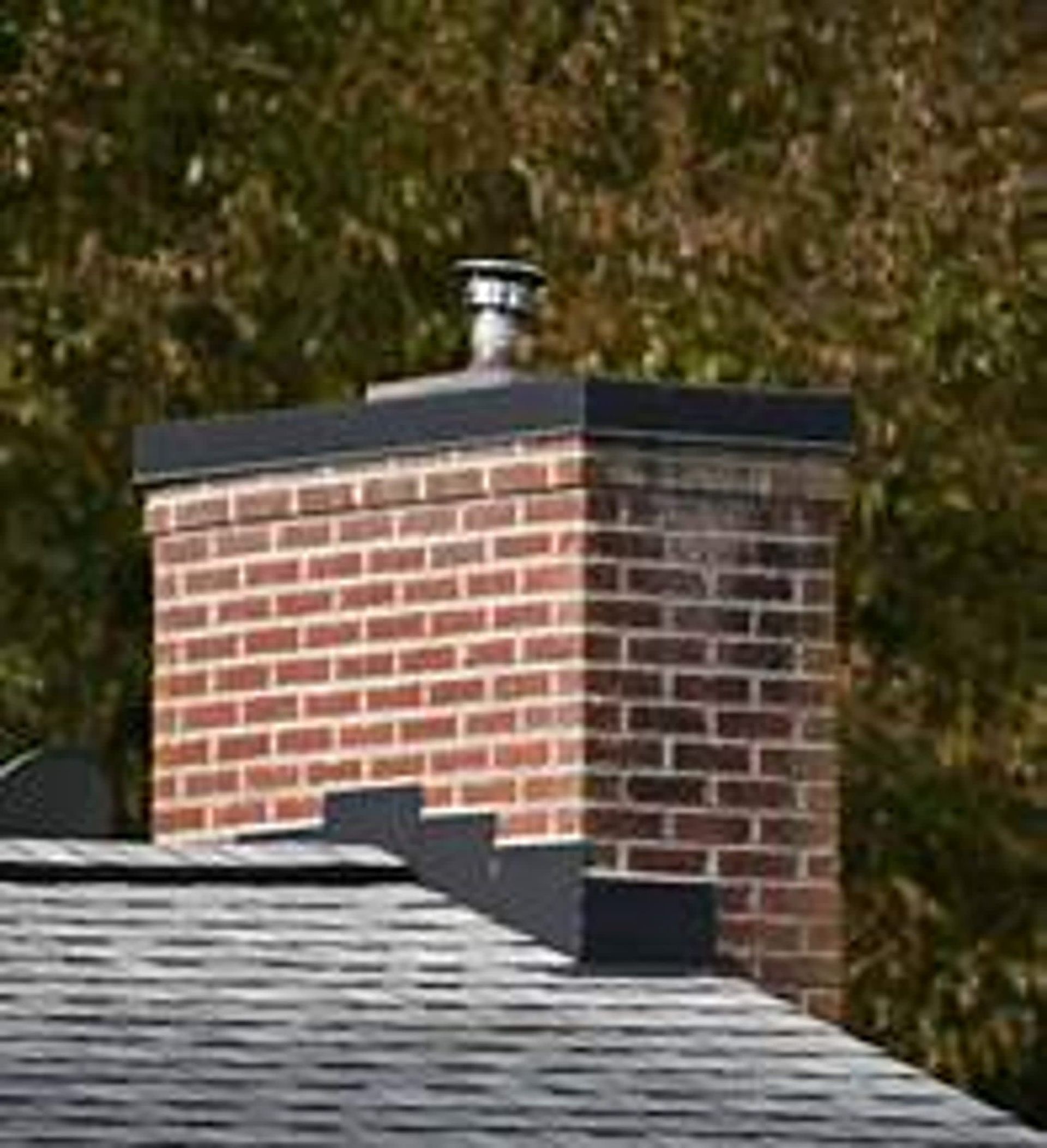 Brick chimney on a shingled roof with a metal cap and flashing, surrounded by trees.