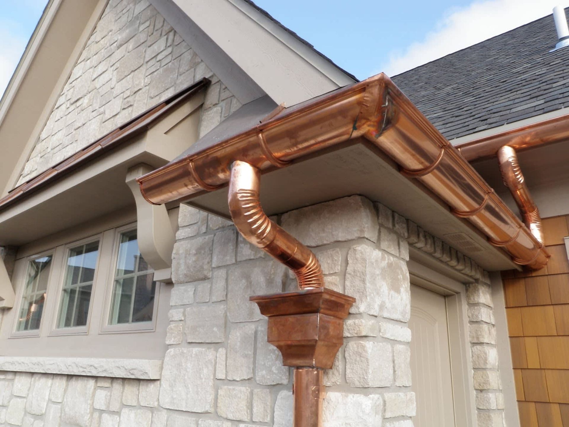 Copper gutters and downspouts on a light stone and wood-clad house.