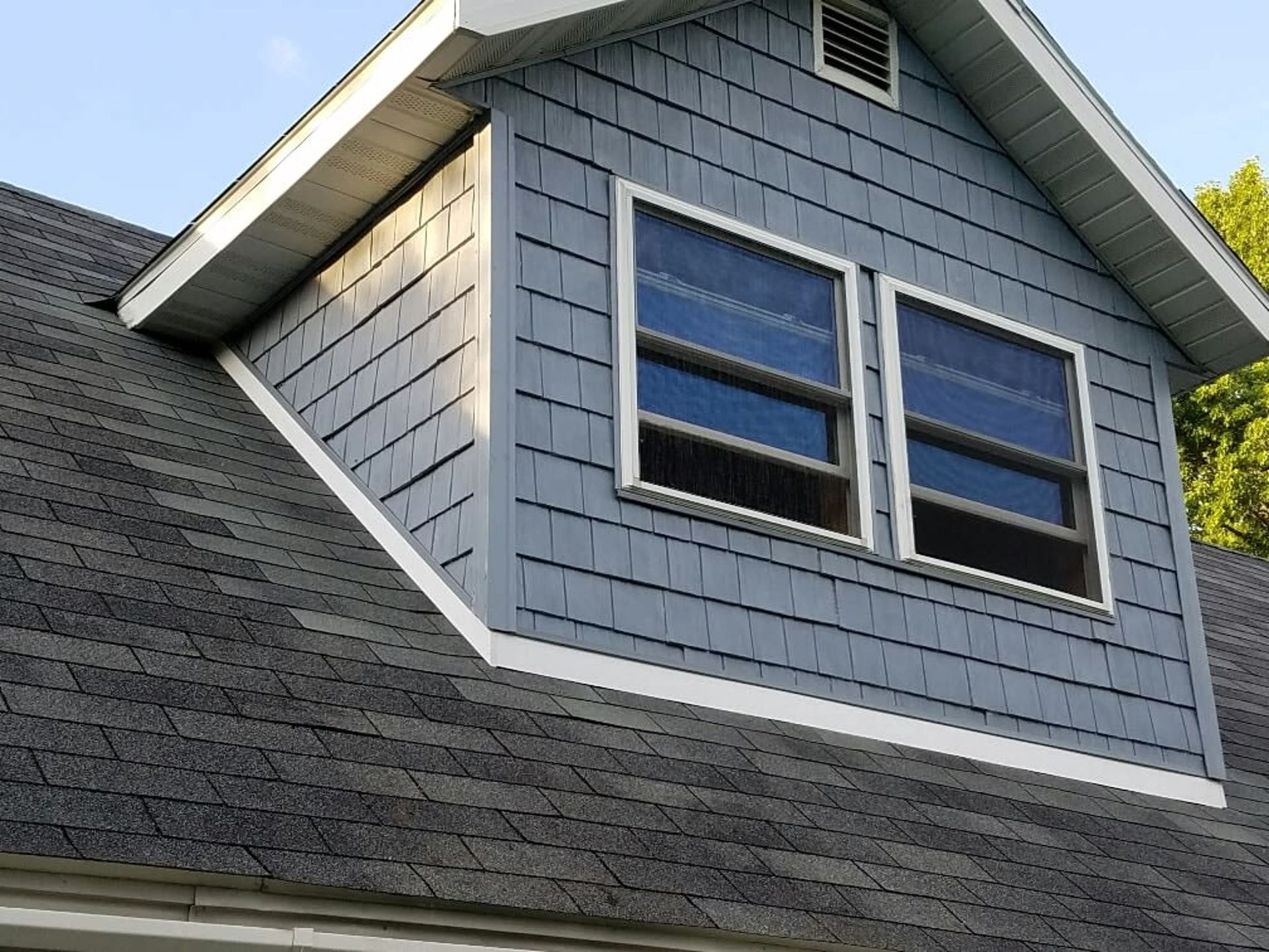 Blue-gray shingle-sided dormer with two windows on a dark gray asphalt shingle roof. White trim and vent.