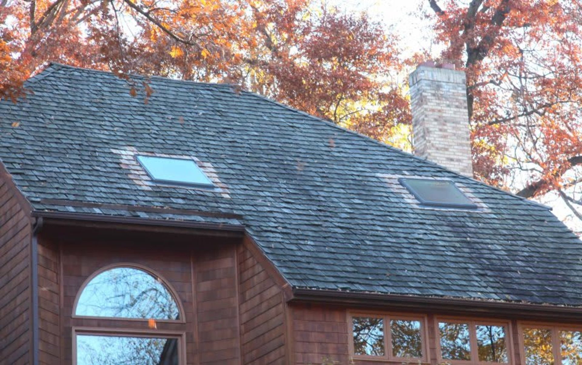 Dark gray shingled roof with two skylights, a brick chimney, and autumn foliage in the background.
