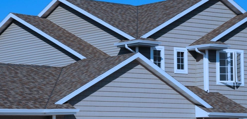 View of a house's complex roof with gray shingles, white trim, and light blue siding against a clear sky.